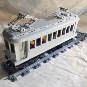 Puede incluir: Un tren de juguete gris con techo blanco y ruedas negras. El tren tiene una fila de ventanas a cada lado y está sobre una vía de plástico gris.