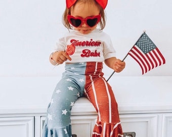 May include: A child's outfit with a patriotic theme. The outfit includes a white t-shirt with the text "American Babe", heart-shaped sunglasses, and bell-bottom pants with a red, white, and blue design. The child is holding a lollipop and an American flag.