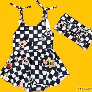 May include: A black and white chequered romper with cartoon character graphics, alongside a matching headband. The romper has a flared skirt and adjustable straps. Graphics include Mickey Mouse, Donald Duck, and Goofy.