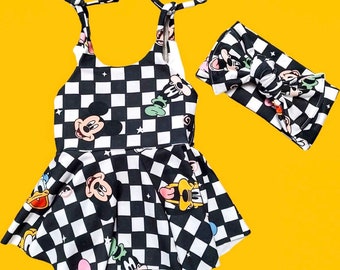 May include: A black and white chequered romper with cartoon character graphics, alongside a matching headband. The romper has a flared skirt and adjustable straps. Graphics include Mickey Mouse, Donald Duck, and Goofy.