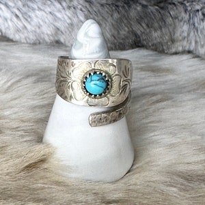 Puede incluir: Un anillo de plata con una piedra turquesa engastada en un diseño floral. El anillo está en un expositor blanco.