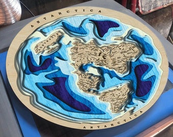 Puede incluir: Un mapa del mundo circular de madera con un océano azul y morado y una masa de tierra beige. El mapa está titulado "ANTARCTICA" y está rodeado por un marco azul.