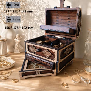 Peut inclure: Une boîte à bijoux en bois brun foncé avec un couvercle à charnière et deux tiroirs coulissants. La boîte présente des bordures en bois clair et des découpes décoratives. Le texte indique des dimensions de 117 x 131 x 143 mm et 156 x 178 x 192 mm.