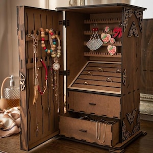 May include: A wooden jewelry armoire with an open door revealing necklaces, bracelets, and earrings. The armoire has multiple compartments, including drawers and earring holders. The wood has a dark brown finish with decorative cutouts.