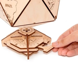 Op de afbeelding: Een houten globe met een kompasvoet. De globe is gemaakt van licht hout en heeft een gedrukte wereldkaart. De kompasvoet is gemaakt van donker hout en heeft een kompasroos. De globe wordt vastgehouden door een hand.