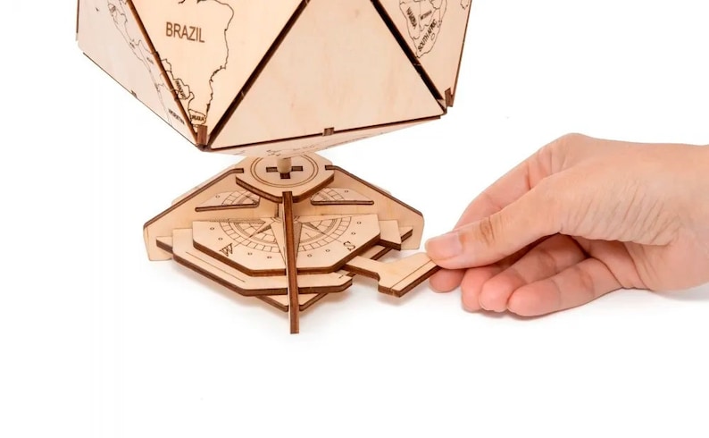 Op de afbeelding: Een houten globe met een kompasvoet. De globe is gemaakt van licht hout en heeft een gedrukte wereldkaart. De kompasvoet is gemaakt van donker hout en heeft een kompasroos. De globe wordt vastgehouden door een hand.