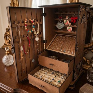 May include: A wooden jewelry armoire with multiple compartments and drawers. The open armoire displays necklaces, bracelets, earrings, and rings. The interior is lined with beige fabric. The armoire is on a wooden dresser.