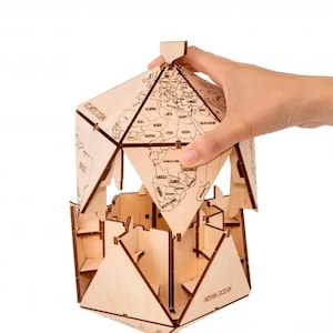 Op de afbeelding: Een houten globe-puzzel met een wereldkaartontwerp. De globe is gemaakt van meerdere stukken die in elkaar passen om een bol te vormen. De globe is gedeeltelijk geassembleerd, met &eacute;&eacute;n stuk dat op zijn plaats wordt gehouden door een hand.