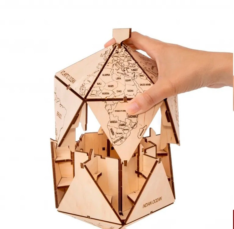Op de afbeelding: Een houten globe-puzzel met een wereldkaartontwerp. De globe is gemaakt van meerdere stukken die in elkaar passen om een bol te vormen. De globe is gedeeltelijk geassembleerd, met &eacute;&eacute;n stuk dat op zijn plaats wordt gehouden door een hand.