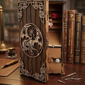 May include: A wooden jewelry box with a dragon design. The box is made of dark brown wood with a laser-cut dragon in a circle on the front. The box is open, revealing compartments for jewelry. The box is on a wooden desk.