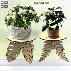 May include: Two wooden plant stands, each with a round top and a decorative leaf-shaped base. One stand holds a gray textured pot with white flowers, the other holds a brown pot with white flowers. The stands are 135 x 250 mm.
