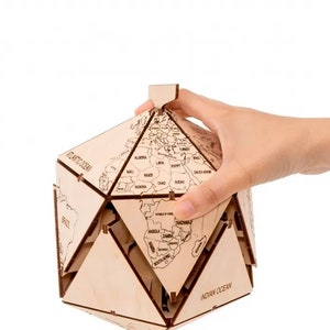 Op de afbeelding: Een houten globe-puzzel met een lichtbruine afwerking. De globe is in een geometrische vorm geassembleerd met de continenten en oceanen met labels. De globe wordt vastgehouden door een hand.