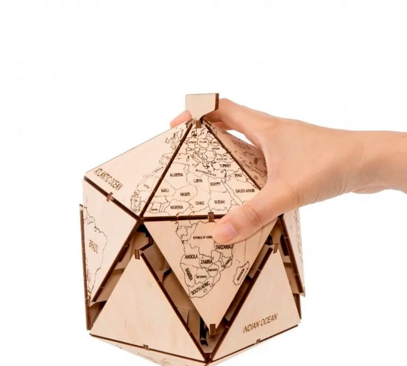 Op de afbeelding: Een houten globe-puzzel met een lichtbruine afwerking. De globe is in een geometrische vorm geassembleerd met de continenten en oceanen met labels. De globe wordt vastgehouden door een hand.