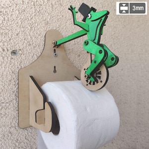 May include: A novelty toilet roll holder with a green frog on a unicycle. The frog, wearing a black top hat, is attached to a wooden backing. The holder is designed to hold a roll of white toilet paper. The holder's thickness is 3mm.