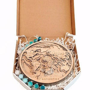 May include: A wooden world map with raised continents and country names, in a cardboard box with a white interior. The box is filled with white shredded paper and contains a set of blue paint pots and a paintbrush.