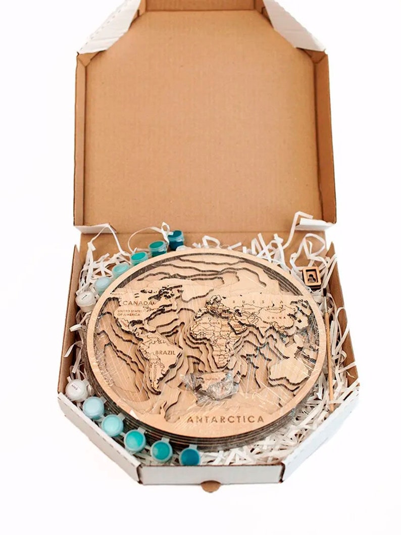 May include: A wooden world map with raised continents and country names, in a cardboard box with a white interior. The box is filled with white shredded paper and contains a set of blue paint pots and a paintbrush.