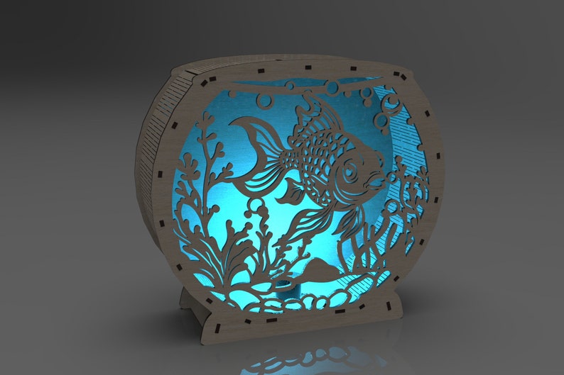 May include: A wooden cutout of a fish tank with a blue light shining through the cutout of a fish and plants. The fish tank is made of wood and has a round shape.