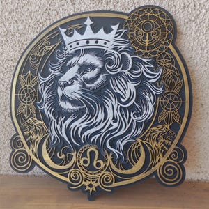 Puede incluir: Una pieza de arte mural de madera en capas que presenta la cabeza de un león con una corona. El diseño incorpora detalles dorados y blancos sobre un fondo gris oscuro. La obra de arte incluye símbolos del zodiaco y patrones decorativos.