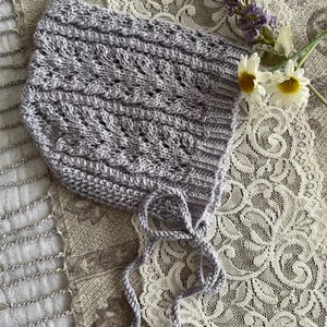 May include: A light grey knitted bonnet with a textured pattern and tie closures. The bonnet is displayed on a lace surface, with sprigs of lavender and daisies. The bonnet is a soft, handmade item.