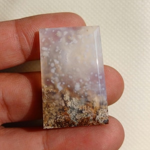 May include: A rectangular polished gemstone cabochon featuring a translucent, milky white upper section with scattered white inclusions and a dense, textured base layer of earthy brown and tan moss-like dendritic patterns throughout the stone.
