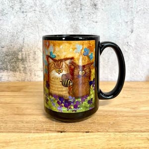 May include: A black ceramic mug featuring a colorful illustration of two fluffy cats, one orange tabby and one brown, with a small bee hovering between them. The scene includes purple flowers and blue butterflies, with the text The Cranky Cat Collection.
