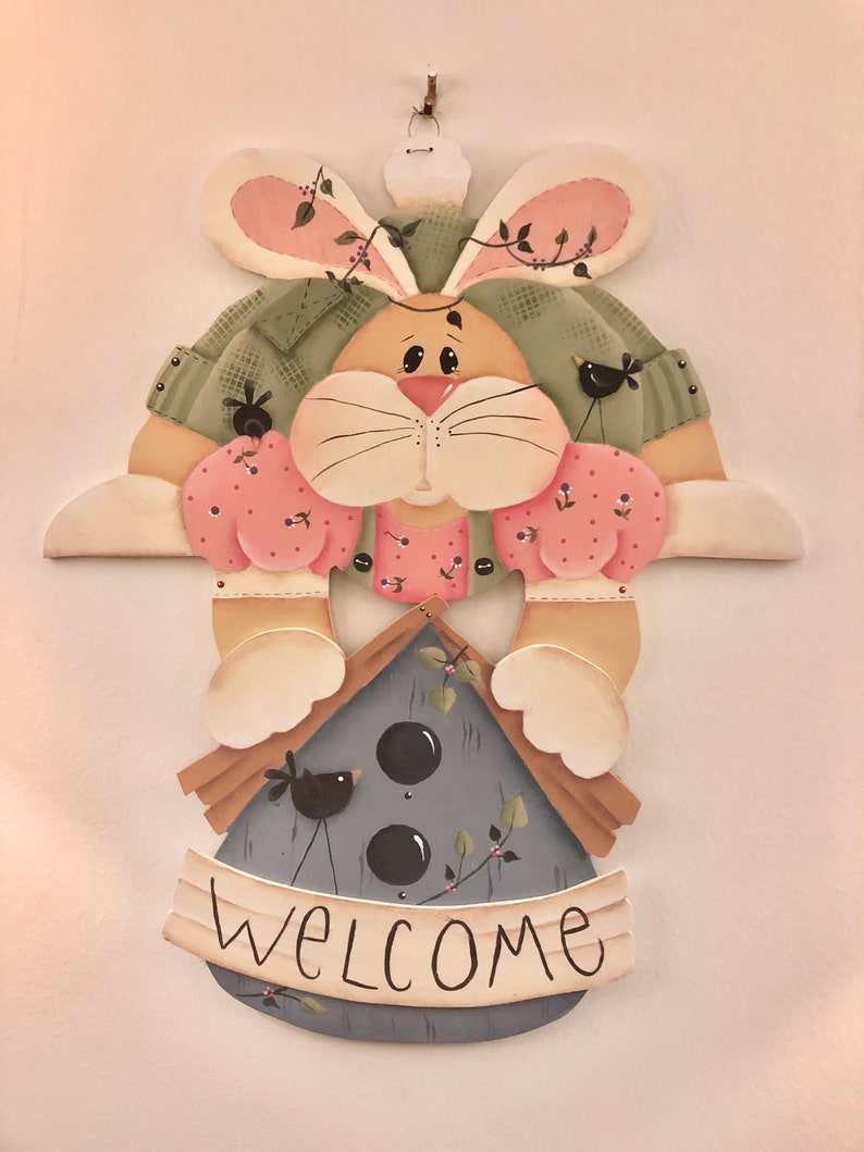 K&ouml;nnte beinhalten: Ein bemalter Holzhase h&auml;lt ein Vogelhaus mit dem Wort "WELCOME" auf einem Banner. Der Hase tr&auml;gt ein rosa-gr&uuml;nes Kleid mit schwarzen V&ouml;geln an den &Auml;rmeln. Das Vogelhaus ist blau mit schwarzen V&ouml;geln und schwarzen Punkten.