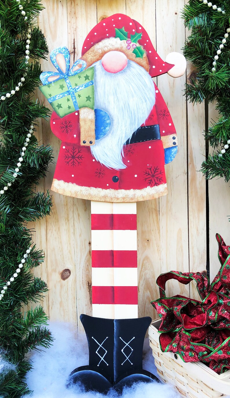 Puede incluir: Una figura de gnomo de Navidad de madera con un gorro a rayas rojas y blancas, pantalones a rayas rojas y blancas y botas negras. El gnomo sostiene una caja de regalo verde con un lazo azul. El gnomo est&aacute; de pie frente a un fondo de madera.