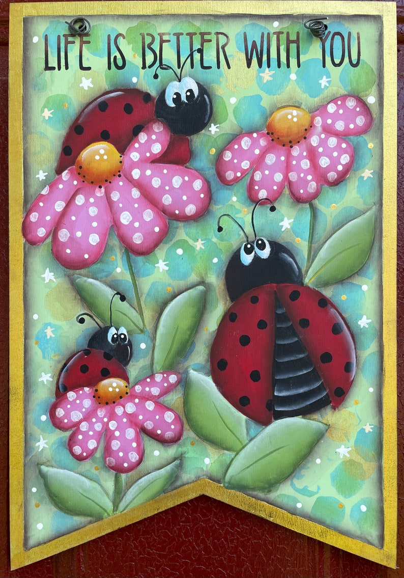 Puede incluir: Un letrero de madera con un borde dorado y el texto "Life is better with you". El letrero est&aacute; decorado con tres mariquitas y flores rosas con lunares blancos sobre un fondo verde.