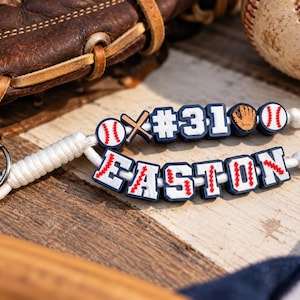 May include: A custom keychain featuring white paracord and a metal ring, adorned with navy blue and white beads. The beads display the name EASTON, the number #31, a baseball, a baseball bat, and a glove, all with red stitching details.