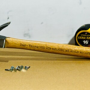 May include: A wooden-handled hammer with a black metal head. The hammer has the words "STANLEY 10 oz" on the side of the head. The handle is engraved with the message "DAD - THANK YOU FOR HELPING ME BUILD MY LIFE!" A yellow tape measure with the words "STANLEY FATMAX 16'" is in the background.