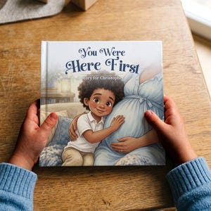 May include: A square hardback book titled You Were Here First: A Story for Christopher. The cover features an illustration of a young child with curly hair hugging the pregnant belly of a person wearing a light blue dress, set in a cosy room.