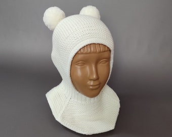 May include: A white knitted balaclava-style hat with two pom-poms on top. The hat is displayed on a mannequin head against a gray background. The hat also includes a neck warmer.