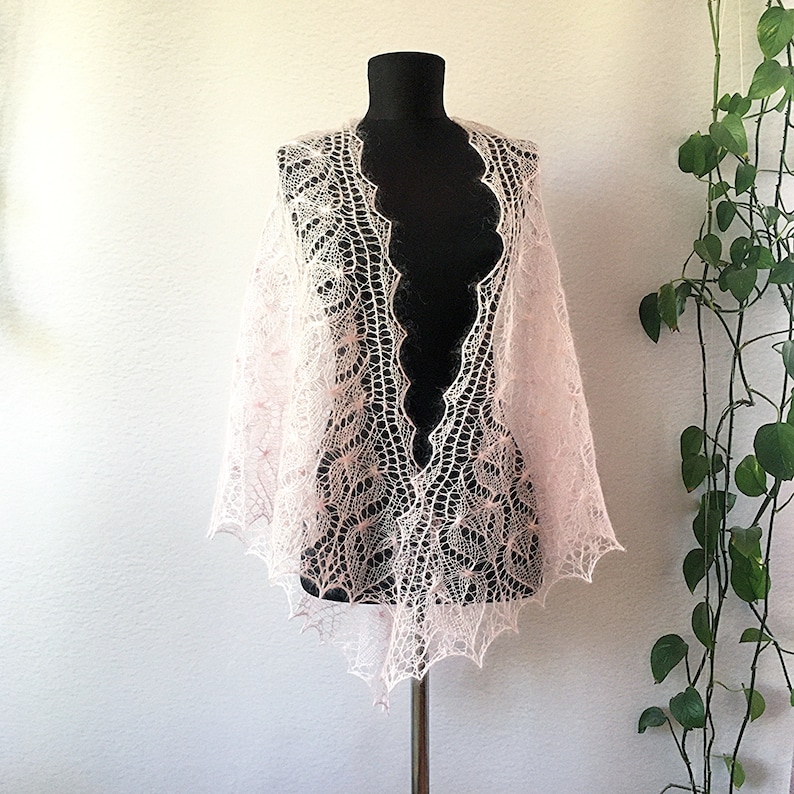 K&ouml;nnte beinhalten: Ein zartes, rosafarbenes gestricktes Tuch mit einem Spitzenmuster. Das Tuch ist &uuml;ber eine schwarze Schaufensterpuppe gelegt und hat einen gewellten Rand.