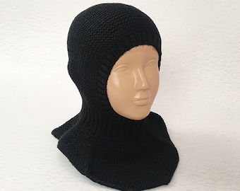 May include: A black knitted balaclava displayed on a mannequin head. The balaclava covers the head, neck, and upper chest, with a close-knit texture. The garment is a solid black color, providing warmth and protection from the elements.