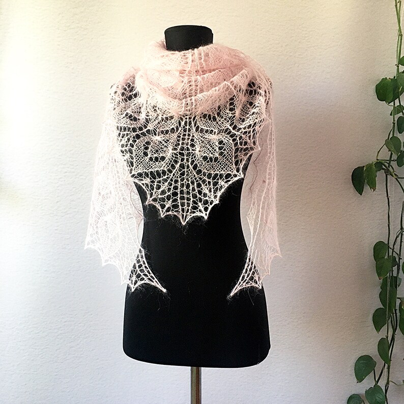 K&ouml;nnte beinhalten: Ein zartes, rosafarbenes gestricktes Tuch mit einem Spitzenmuster, das auf einer schwarzen Schaufensterpuppe pr&auml;sentiert wird.