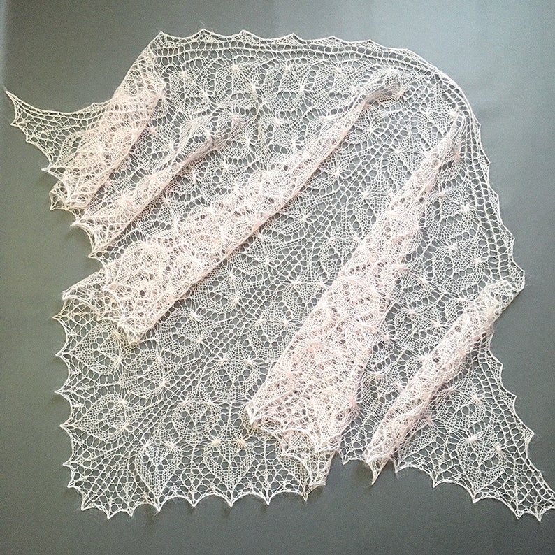 K&ouml;nnte beinhalten: Ein zartes, handgestricktes Tuch in einem sanften Rosa- und Wei&szlig;ton. Das Tuch ist aus feinem Garn gefertigt und hat ein zartes, durchbrochenes Muster.