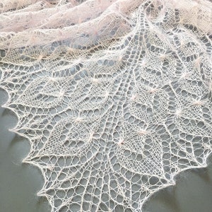K&ouml;nnte beinhalten: Ein zartes, blassrosa gestricktes Tuch mit einem Spitzenmuster. Das Tuch ist aus feinem Garn gefertigt und hat eine weiche, flie&szlig;ende Textur.