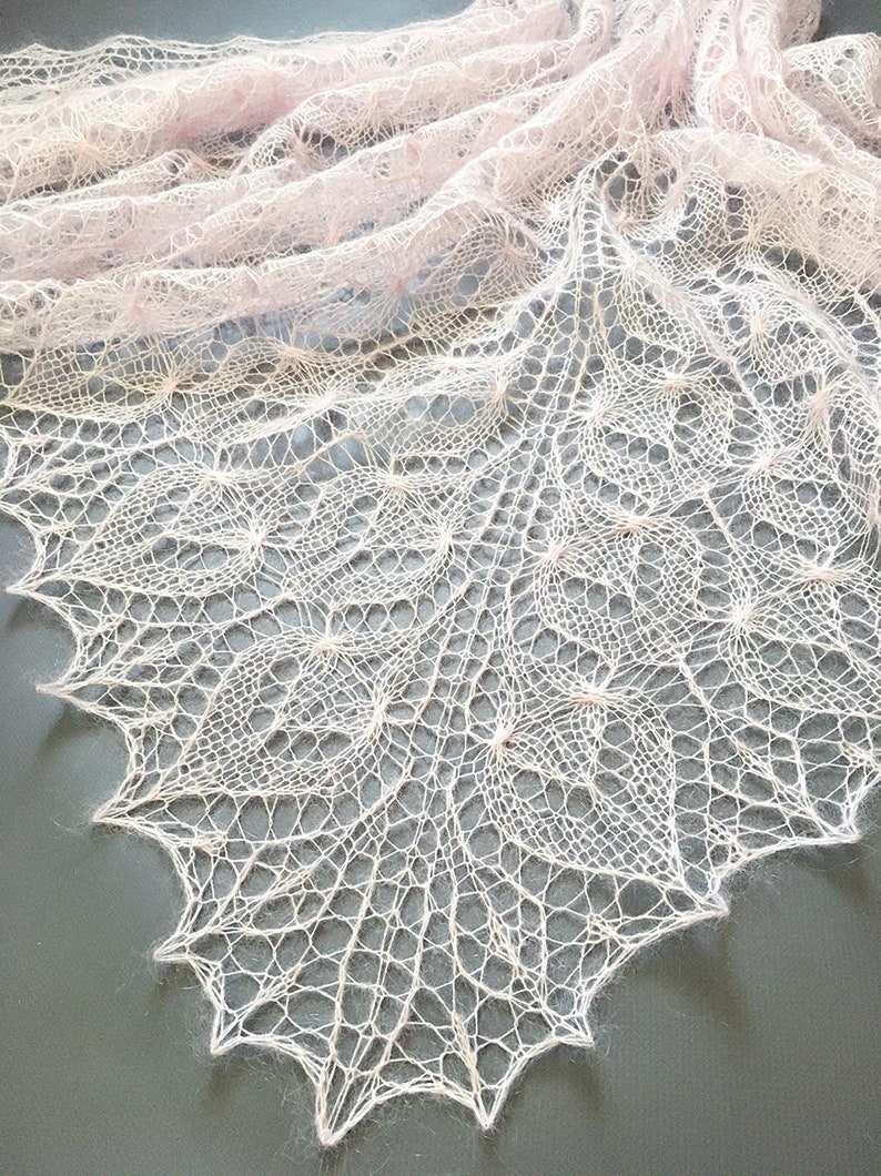 K&ouml;nnte beinhalten: Ein zartes, blassrosa gestricktes Tuch mit einem Spitzenmuster. Das Tuch ist aus feinem Garn gefertigt und hat eine weiche, flie&szlig;ende Textur.