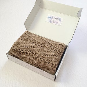K&ouml;nnte beinhalten: Ein beigefarbener Strick Schal mit einem zarten Spitzenmuster ist gefaltet und in einer wei&szlig;en Schachtel mit geometrischem Muster verpackt. Die Schachtel tr&auml;gt ein Etikett mit dem Text "MariKnit" und Kontaktinformationen.