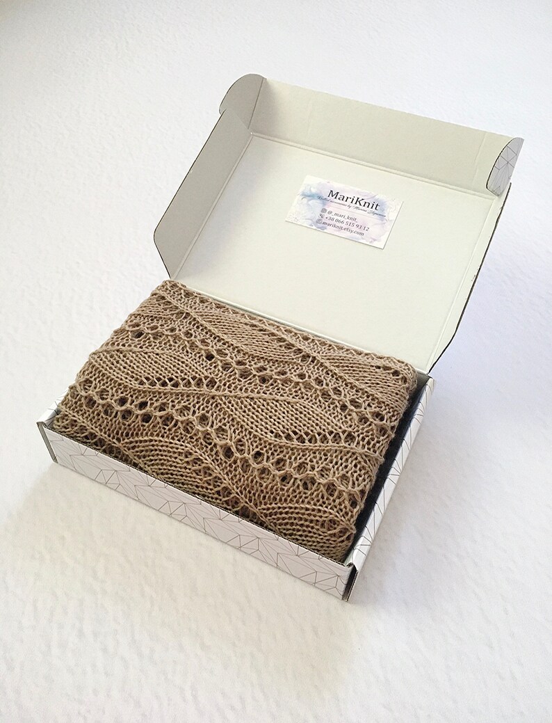 K&ouml;nnte beinhalten: Ein beigefarbener Strick Schal mit einem zarten Spitzenmuster ist gefaltet und in einer wei&szlig;en Schachtel mit geometrischem Muster verpackt. Die Schachtel tr&auml;gt ein Etikett mit dem Text "MariKnit" und Kontaktinformationen.