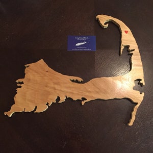 May include: A light brown wooden cutout map of Cape Cod featuring a small red heart icon on the upper peninsula. A rectangular blue business card is positioned near the center, displaying the text Long Island Birch and a website address.