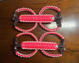 May include: Four pink and white paracord bracelets with black accents and silver metal clasps. The bracelets are connected together in pairs.