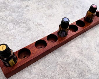 May include: A wooden essential oil rack with eight slots, featuring three bottles of essential oil. The rack is made of dark red wood and has a natural wood grain. The bottles are all glass and have black caps. The bottles are labeled with the word "TERRA" in white lettering.