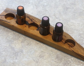 May include: Wooden essential oil holder with five slots. The holder is made of dark wood with a natural finish. Three bottles of essential oil are in the holder. The bottles are labeled "Orange", "Lemon", and "Lavender".
