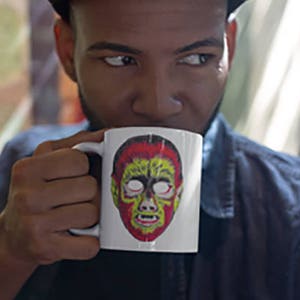Könnte beinhalten: Eine weiße Keramiktasse mit einem bunten Motiv eines Monstergesichts. Die Tasse wird von einer Person gehalten, die eine schwarze Mütze und ein blaues Jeanshemd trägt.