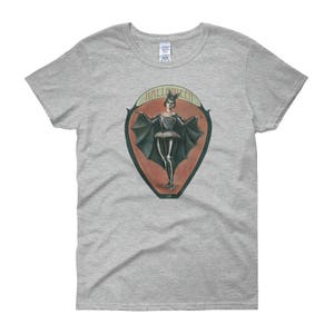 May include: A gray t-shirt with a vintage-style illustration of a woman in a bat costume. The illustration is surrounded by a red and orange oval with the word "HALLOWEEN" printed in black.