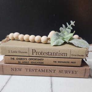 May include: A stack of three brown books with the titles "Protestantism", "The Church's Opportunity in Adult Education", and "New Testament Survey". A wooden bead garland with green foliage is on top of the books.