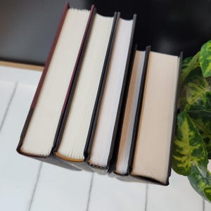 以下が含まれることがあります： 白いページと黒い背表紙を持つハードカバーの本が4冊。左側の本はえんじ色の背表紙です。