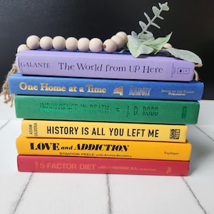 Puede incluir: Una pila de libros coloridos con títulos como "The World from UP Here" y "5-FACTOR DIET". Una guirnalda de cuentas de madera y una ramita de vegetación descansan encima. Los libros son de varios colores, incluyendo morado, azul, verde, amarillo y rojo.