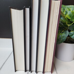 以下が含まれることがあります： 白いページと黒い背表紙を持つハードカバーの本が4冊、白い表面に立てられています。一番右の本は、えんじ色の背表紙です。